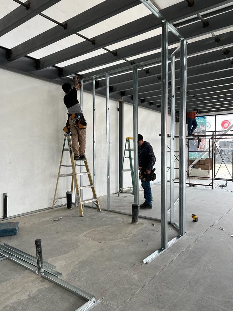 Hombre arriba de una escalera trabajando en la instalacion de postes metálicos, al fondo dos hombres trambien trabajan, los tres en un proyecto comercial de bodega de Kelder Zertuche Ensenada