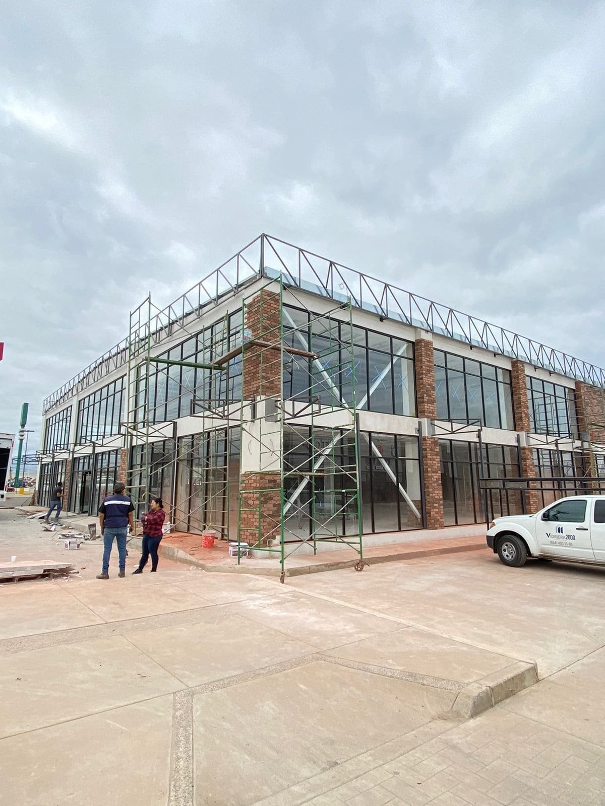 Fachada de edificio comercial moderno de dos niveles. La estructura tiene amplios ventanales con vidrio templado con marcos negros y columnas revestidas de ladrillo. Se observan tambien andamios y dos trabajadores al lado de la entrada principal bajo un cielo nublado.