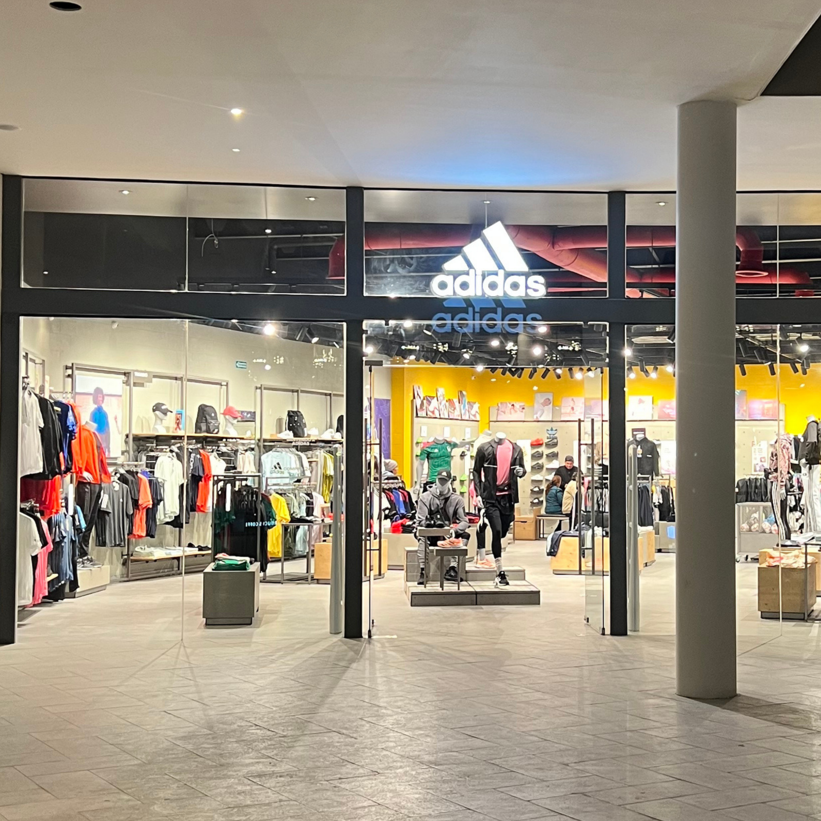 Vista frontal de la entrada de la tienda Adidas en Plaza comercial Península Tijuana, se observan ventanales de vidrio y en el interior hay exhibidores y percheros con ropa deportiva de diversos colores.