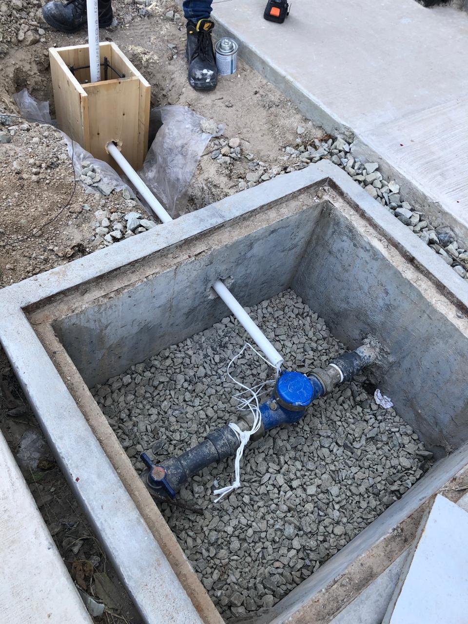 Registro de concreto con fondo de grava que contiene un medidor de agua y tuberías, conectado a una instalación exterior con cimbra de madera en el suelo, en proyecto de sistema hídrico en tijuana